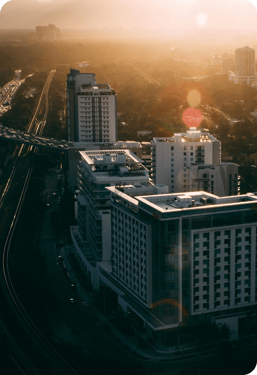 Aerial view of a cityscape at sunset, with rays illuminating modern high-rise buildings and a busy highway. Warm, soft glow creates a serene, urban ambiance.